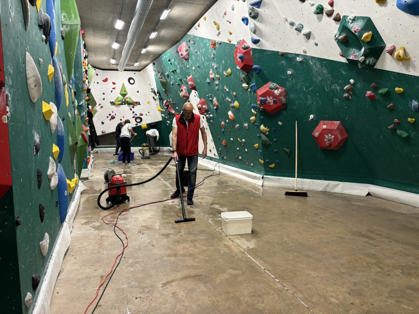 Menschen reinigen den Boden einer Kletterhalle nach einem Wasserschaden mit Staubsauger, Eimer und Besen.